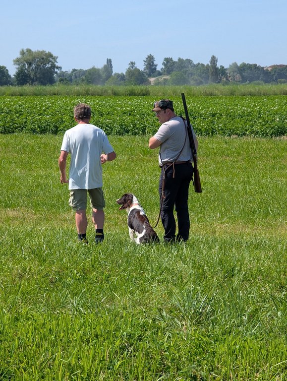 Zwei Männer und ein Hund stehen auf einem grünen Feld. Einer hält ein Gewehr, der Hund sitzt still. Im Hintergrund sind Bäume und blauer Himmel sichtbar.